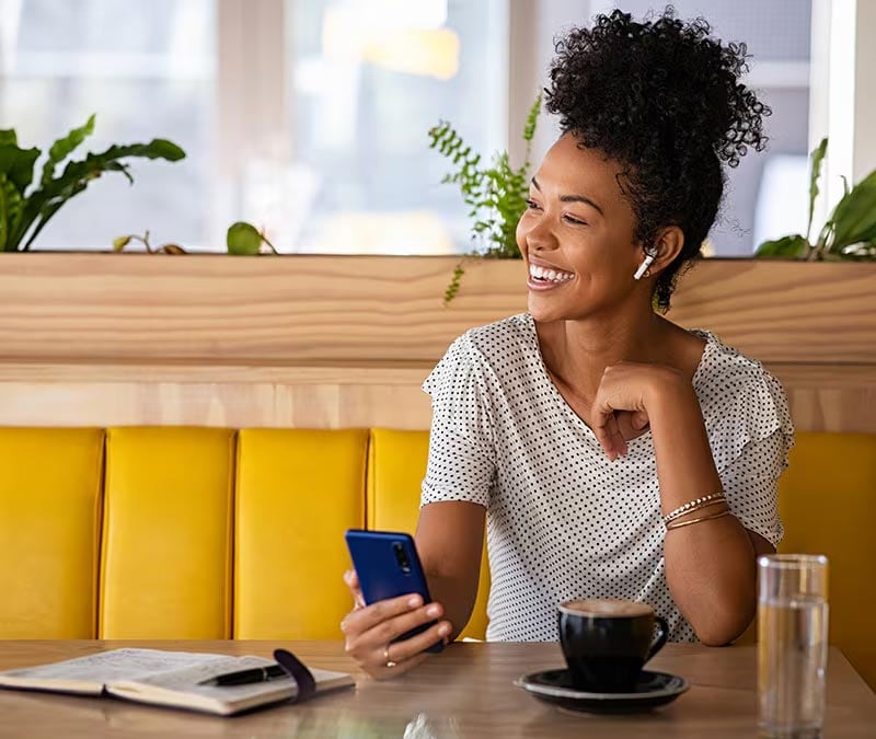 Lächelnde Frau mit iPhone in einem Café, nachdem sie gelernt hat, wie sie durch das Einrichten eines VPN ihr iPhone besser schützen kann.