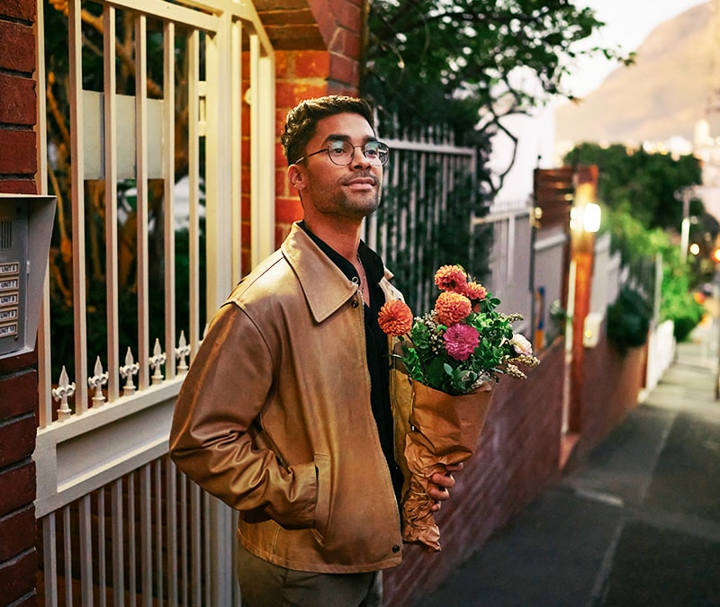 Ein Mann wartet vor einem Backsteinhaus und hält einen Blumenstrauß in der Hand.