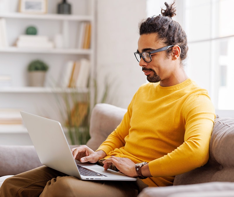 A man in glasses and a yellow shirt sitting on a couch with a laptop, engrossed in his work and wondering whether his Mac can get viruses.