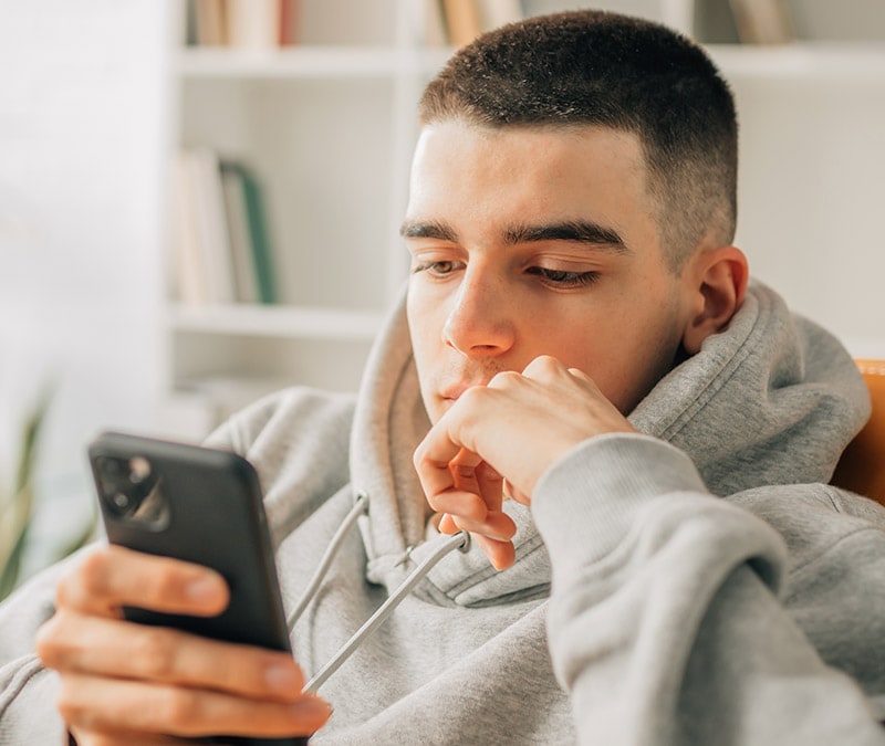 Ein junger Mann in einem grauen Kapuzenpulli studiert aufmerksam sein iPhone und sucht nach Informationen darüber, was iPhone-Viren sind und wie man sie loswerden kann.