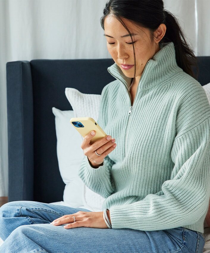 Eine Frau sitzt auf einem Bett, blickt auf ihr Telefon und überlegt, wie sie ein einprägsames Passwort erstellen kann.