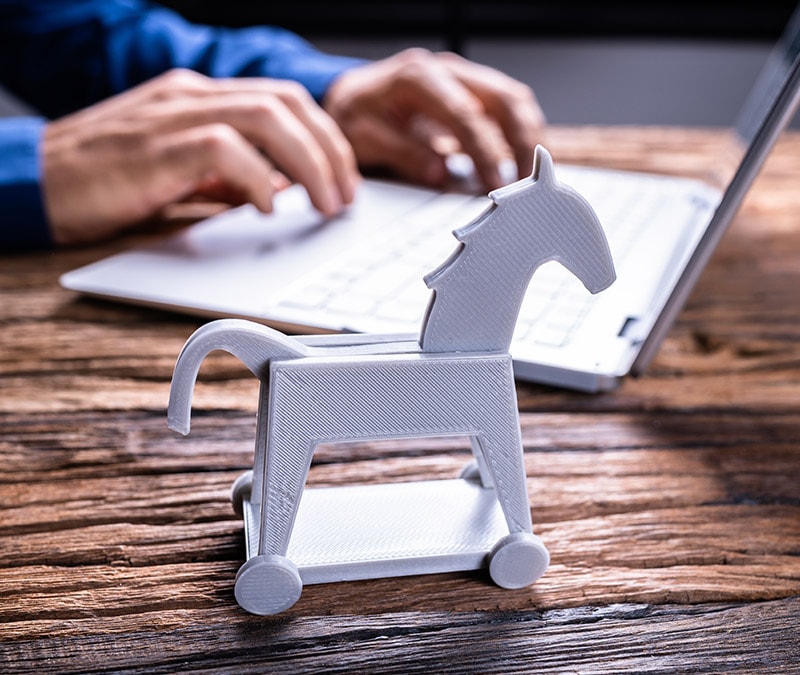 Modell eines trojanischen Pferds auf einem Schreibtisch zur Symbolisierung von Trojanern mit einer Person mit Laptop im Hintergrund zur Veranschaulichung des Themas der Erkennung und Entfernung von Trojanern.